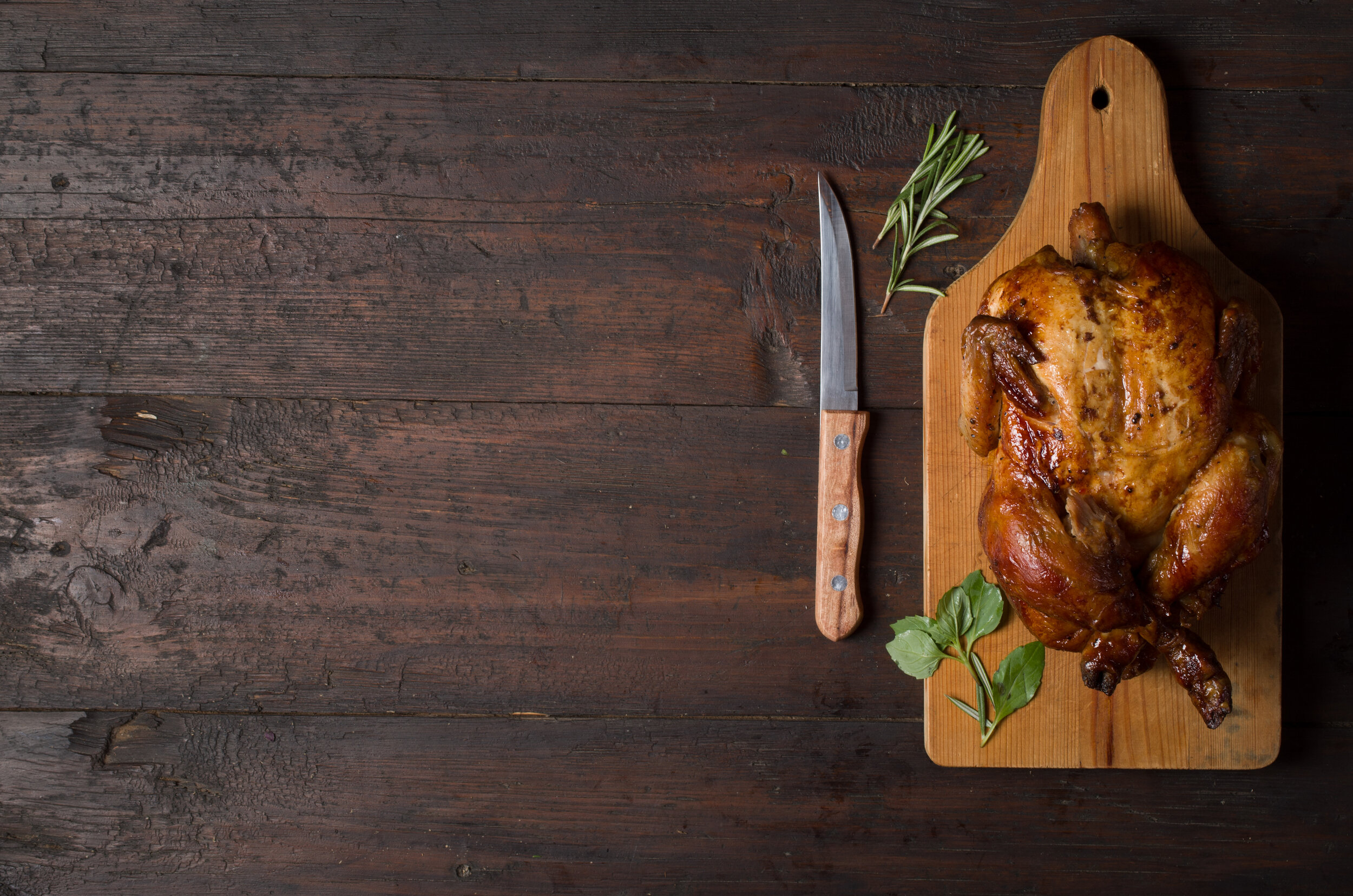 Roasted chicken on a wooden cutting board with a knife and fresh herbs, showcasing a delicious meal preparation scene.