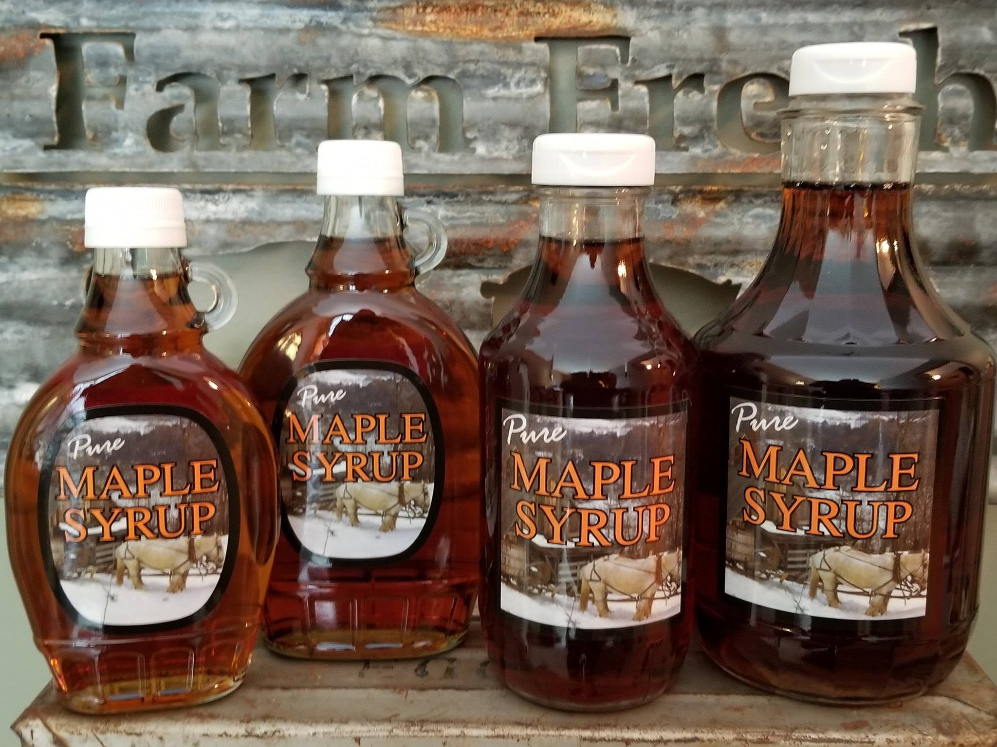 Four bottles of pure maple syrup in various sizes are displayed against a rustic backdrop featuring the words "Farm Fresh." The labels prominently feature the phrase "Pure Maple Syrup," highlighting the product's natural quality.