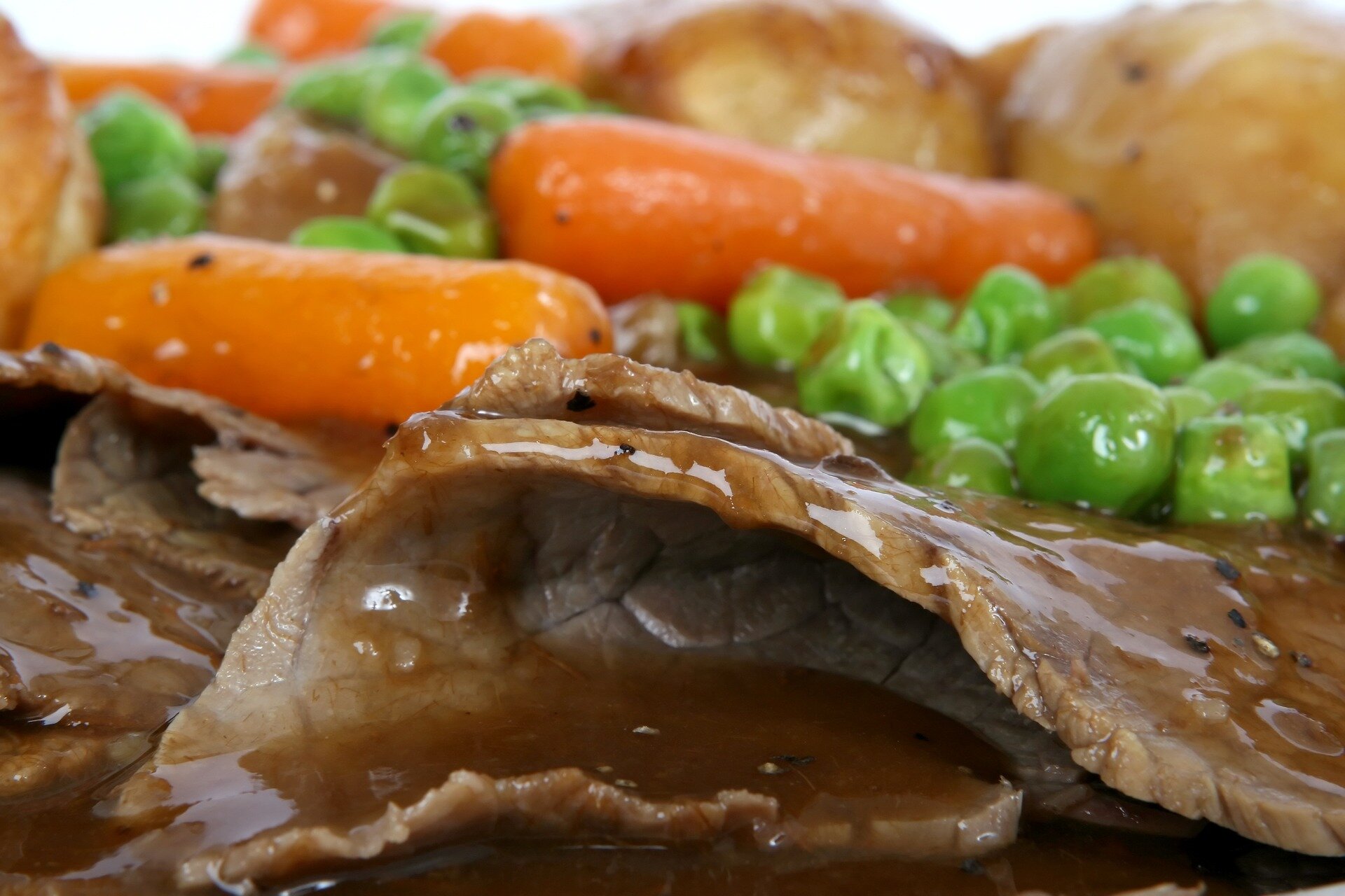 Close-up of tender roast beef slices drizzled with savory gravy, accompanied by vibrant carrots, green peas, and roasted potatoes, showcasing a hearty and appetizing meal.