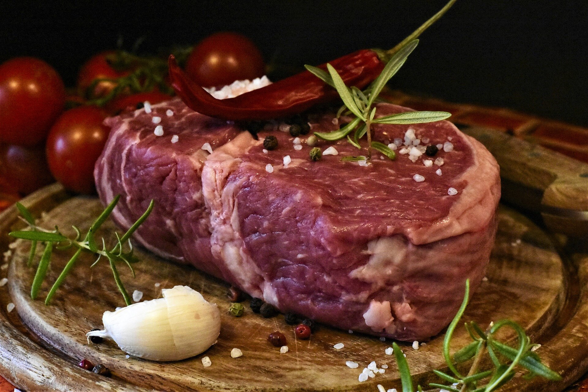 Fresh cut beef steak on a wooden board, garnished with garlic, rosemary, and chili pepper, surrounded by cherry tomatoes and black peppercorns, showcasing a vibrant and appetizing arrangement.
