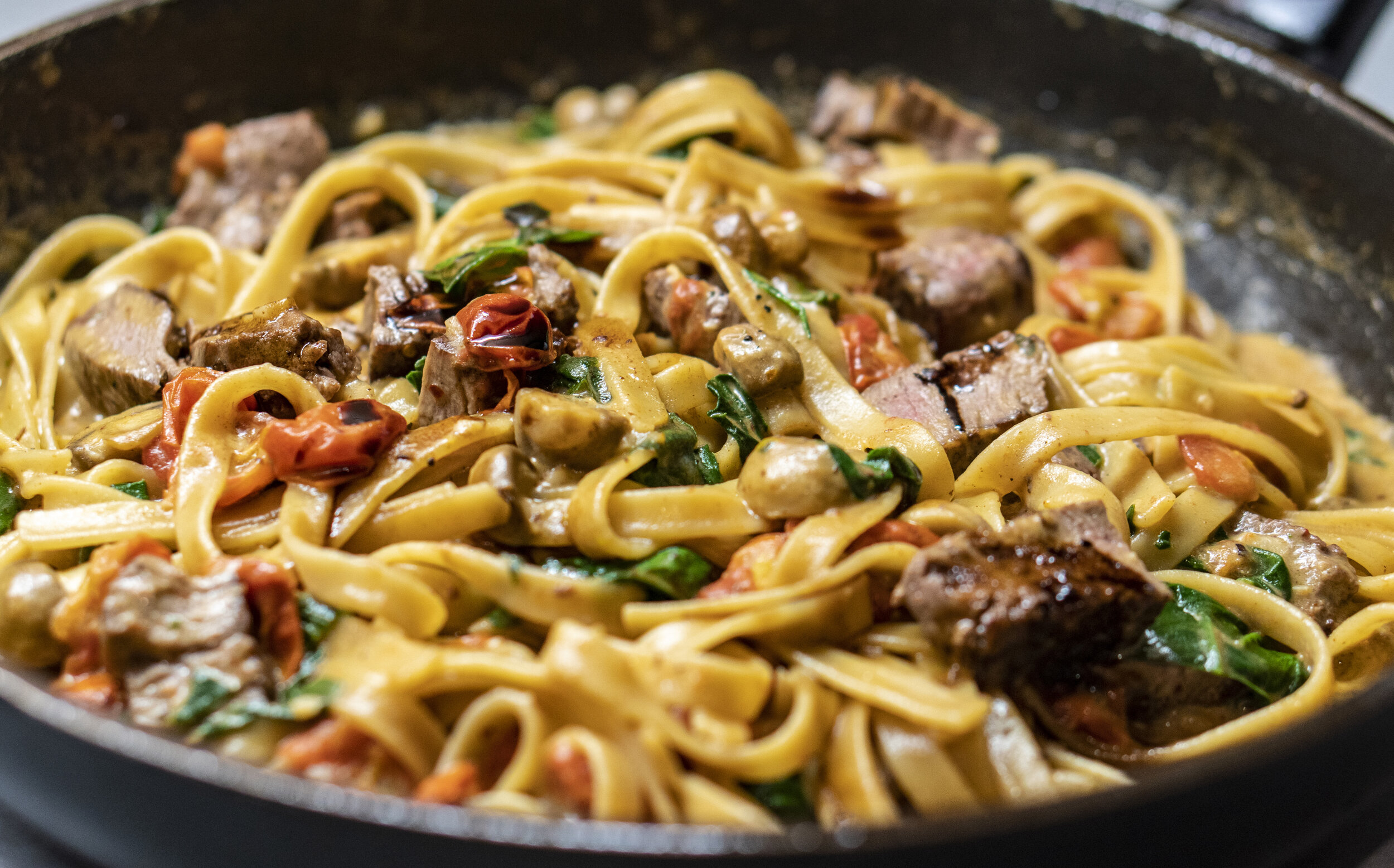 Creamy fettuccine pasta with tender beef, spinach, and sun-dried tomatoes, cooked in a skillet for a delicious and hearty meal.