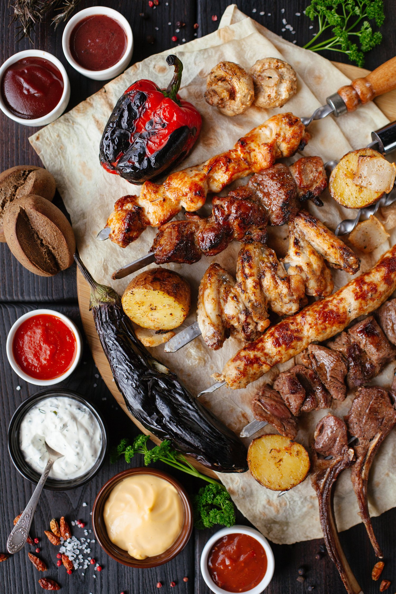image-asset Grilled meat platter featuring assorted skewers of chicken, beef, and lamb, accompanied by roasted vegetables, sauces, and dips. The arrangement includes a charred red pepper, mushrooms, and seasoned potatoes, all presented on parchment paper with fresh herbs and spices. Perfect for barbecue enthusiasts and food lovers.
