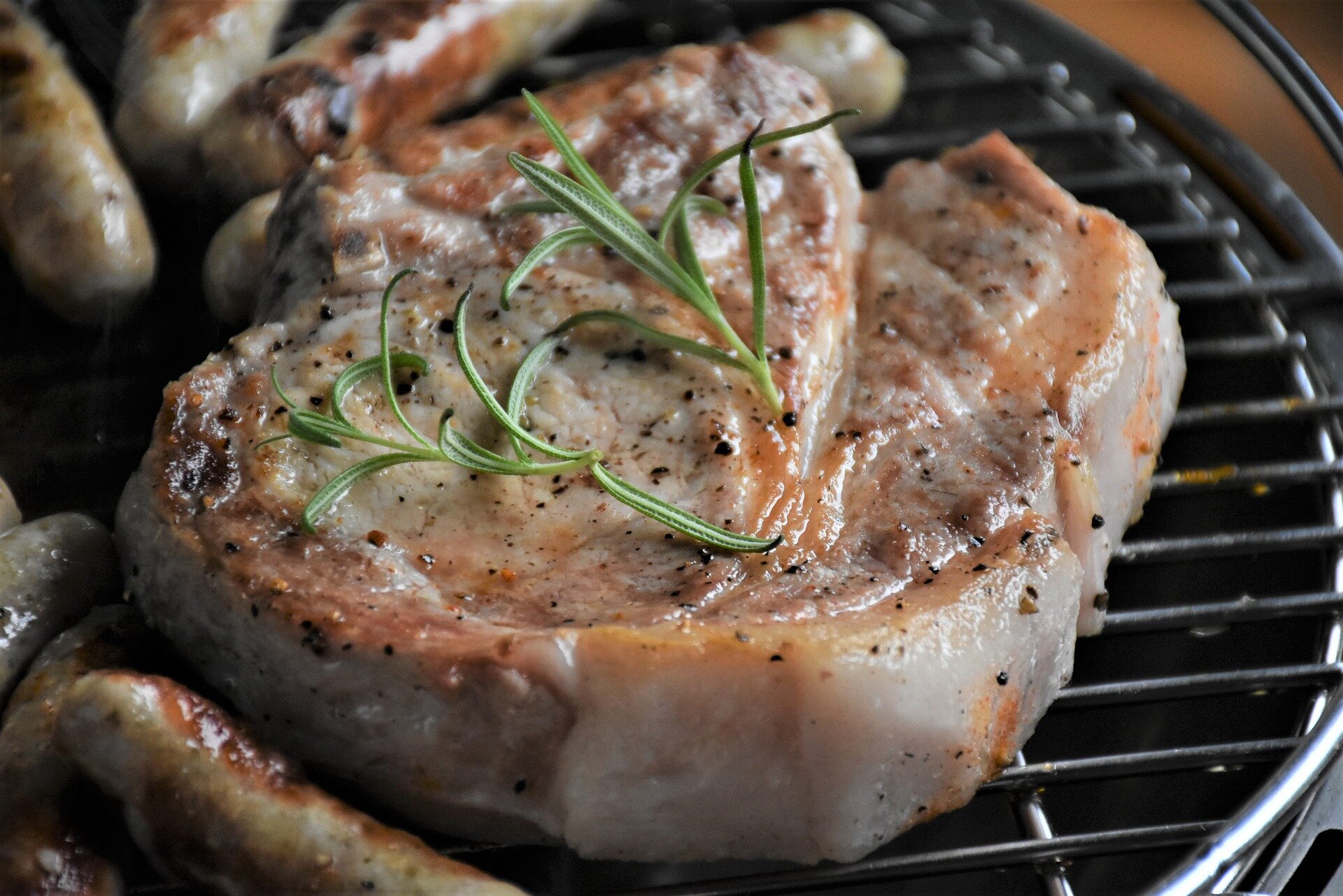 Grilled pork chop with rosemary garnish, surrounded by sausages on a barbecue grill.