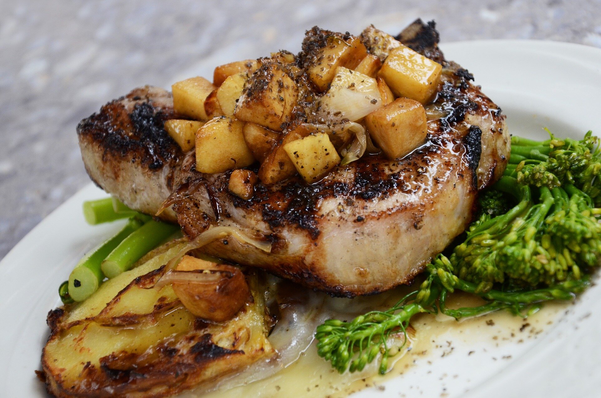 meat-802046_1920 Grilled pork chop topped with caramelized apples and onions, served with roasted potatoes and steamed broccolini on a white plate.