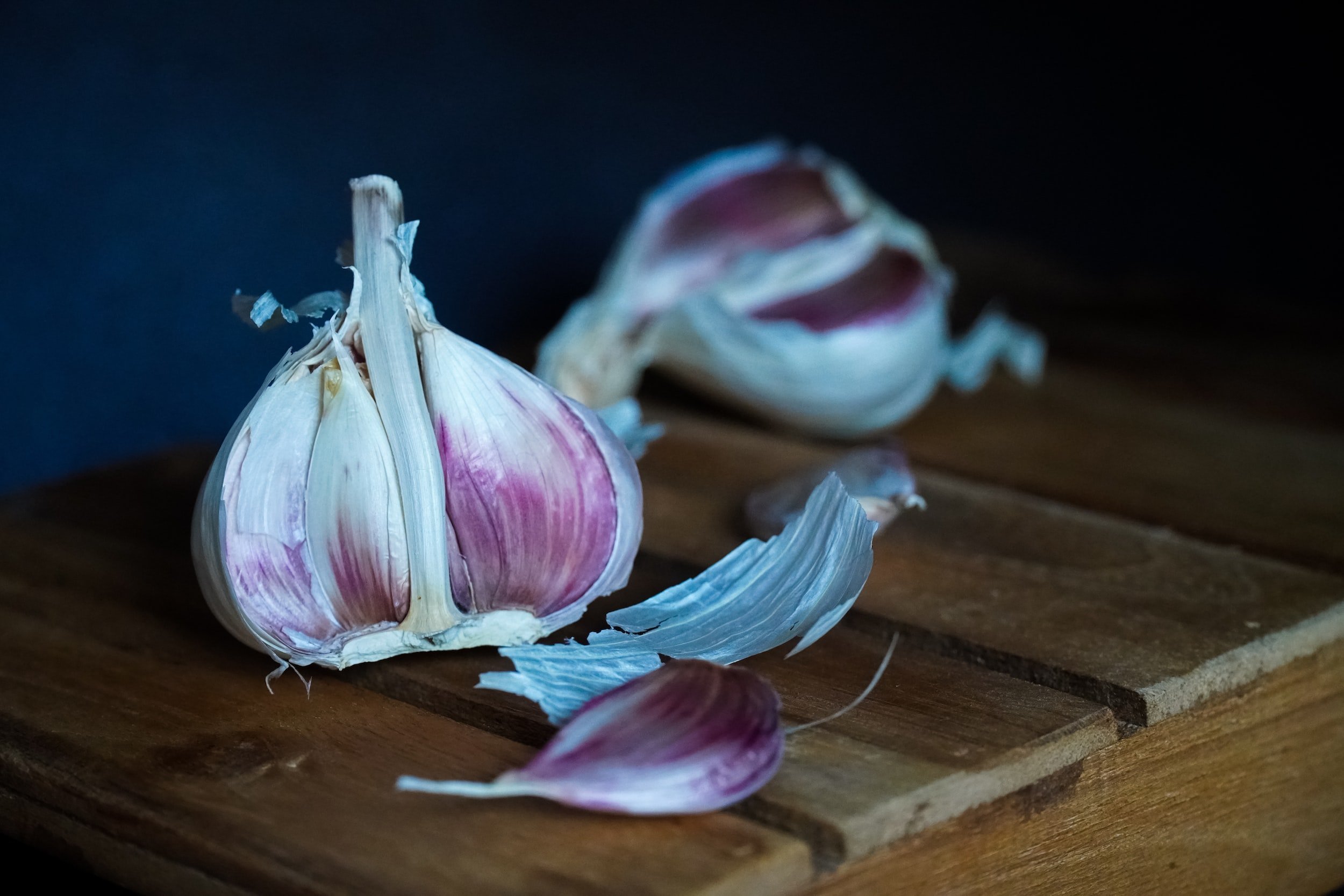unsplash-image-PUFx3691Hgs Fresh garlic bulbs and cloves scattered on a wooden surface, showcasing their purple skin and papery layers against a dark background. Ideal for culinary uses and health benefits.